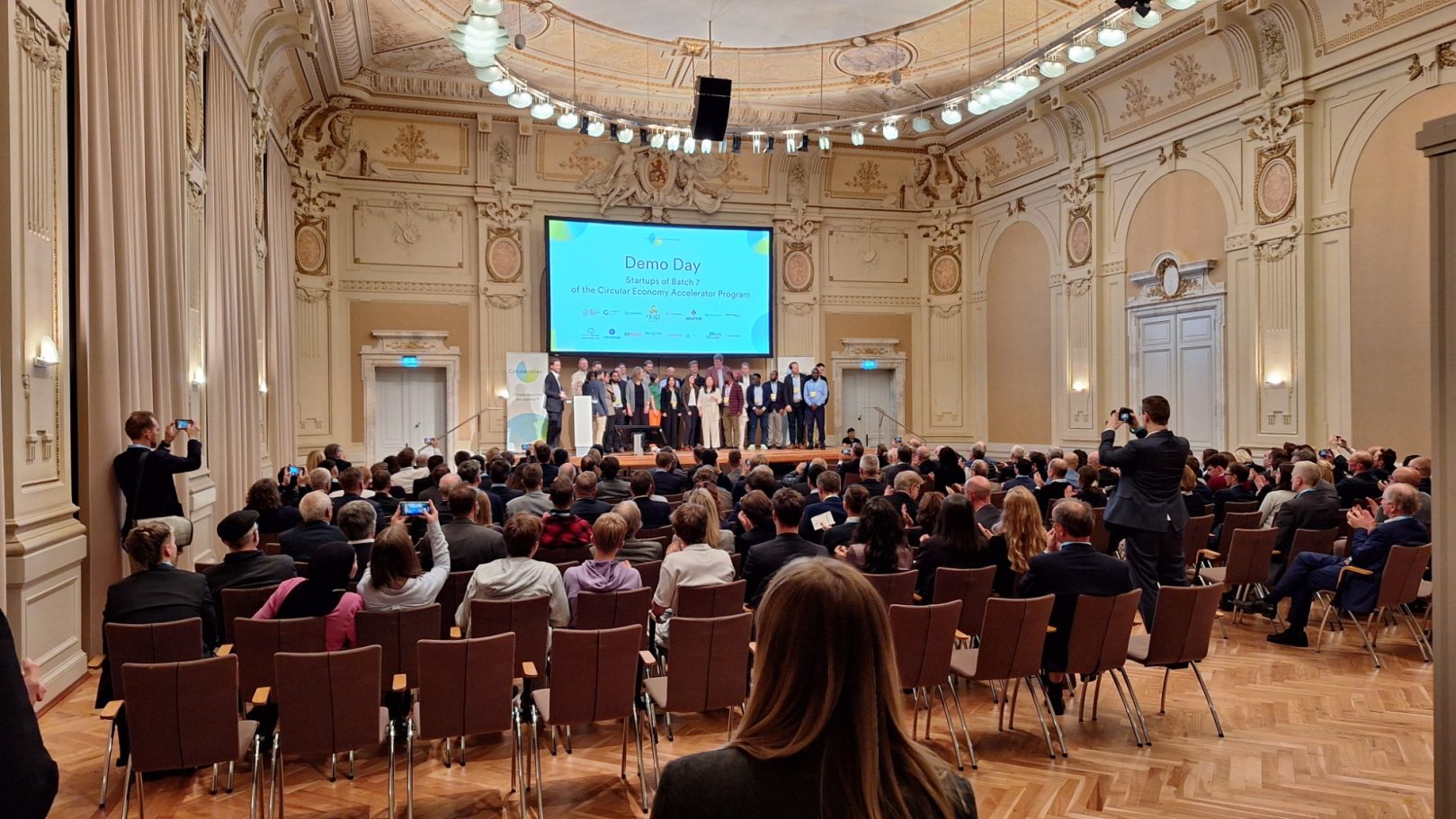 Eine vollbesetzte Stadthalle Wuppertal mit einem Podium und Präsentation.